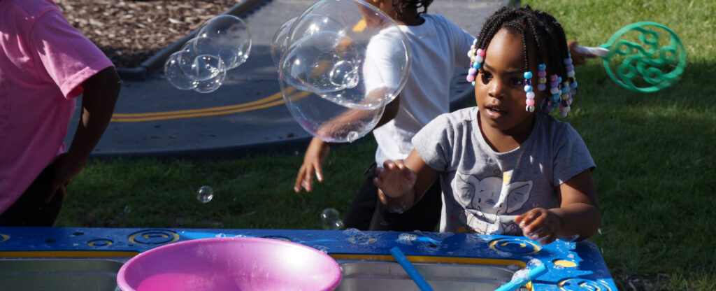 A girl looks in wonder as bubbles fly by her from the bubble table outside in the garden area, called sproutside